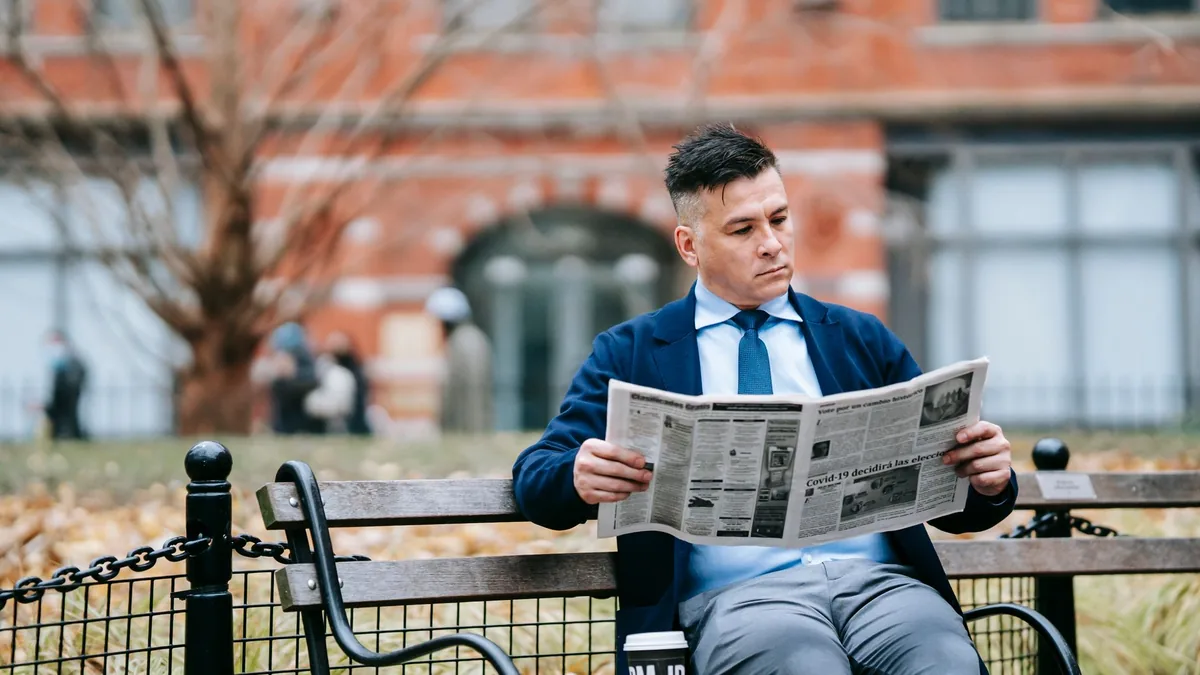 A man in formal attire sits on a park bench reading a ne 6326332